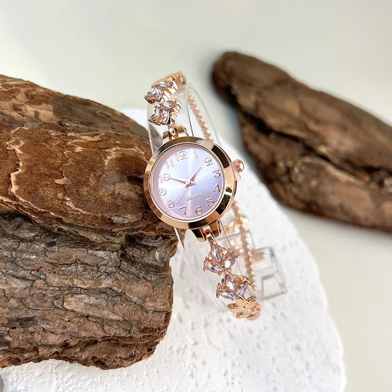 elógio feminino sofisticado, com caixa redonda em liga metálica, mostrador clássico com números árabes e ponteiros em formato de folha. Pulseira delicada com detalhes em strass, combinando charme e estilo