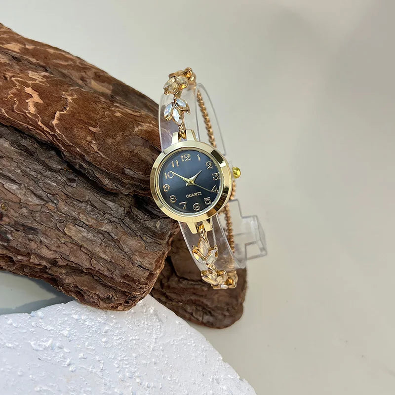 elógio feminino sofisticado, com caixa redonda em liga metálica, mostrador clássico com números árabes e ponteiros em formato de folha. Pulseira delicada com detalhes em strass, combinando charme e estilo