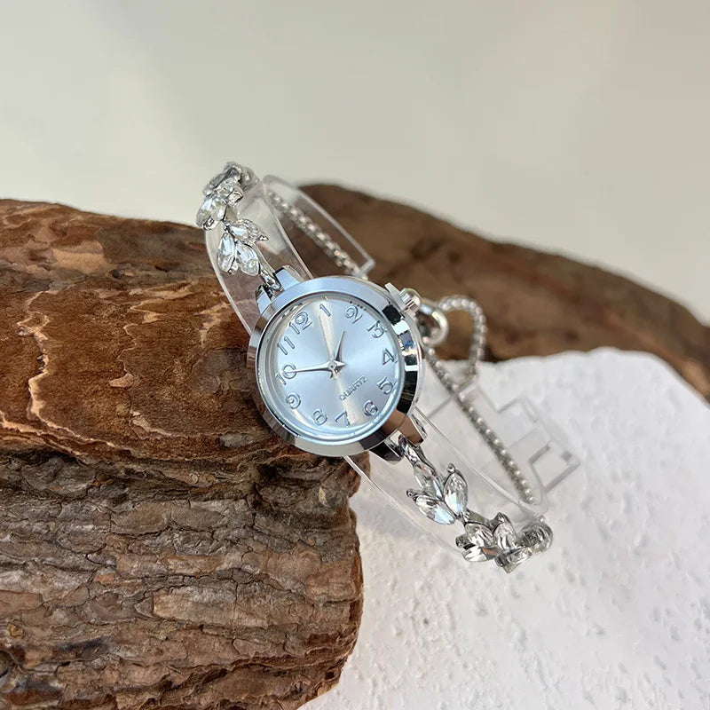 elógio feminino sofisticado, com caixa redonda em liga metálica, mostrador clássico com números árabes e ponteiros em formato de folha. Pulseira delicada com detalhes em strass, combinando charme e estilo