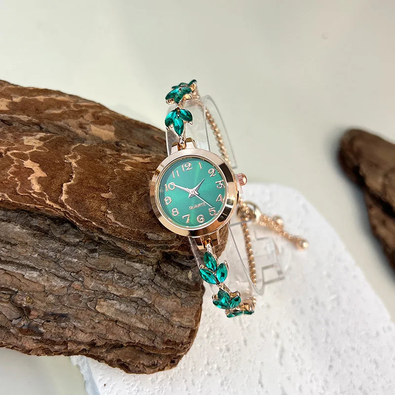 elógio feminino sofisticado, com caixa redonda em liga metálica, mostrador clássico com números árabes e ponteiros em formato de folha. Pulseira delicada com detalhes em strass, combinando charme e estilo