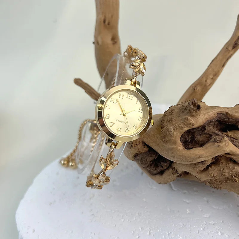 elógio feminino sofisticado, com caixa redonda em liga metálica, mostrador clássico com números árabes e ponteiros em formato de folha. Pulseira delicada com detalhes em strass, combinando charme e estilo